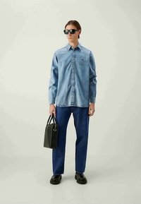 Camisa de ganga em azul claro com fecho de botão, combinada com jeans azul escuro. Uma bolsa e sapatos pretos completam o visual. Óculos de sol pretos estão usados.