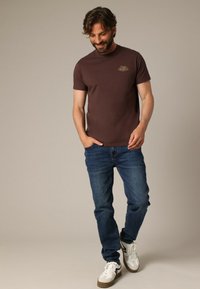 T-shirt en coton marron à manches courtes avec un détail logo. Porté avec un jean bleu et des baskets blanches avec des accents marron.