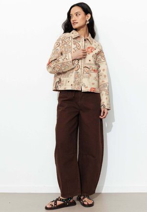 Femme aux longs cheveux foncés porte une veste beige à motif cachemire, un pantalon large marron foncé et des sandales noires cloutées, debout contre un mur blanc.