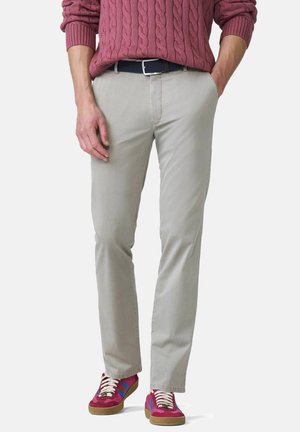 Homme portant un pantalon beige, une ceinture foncée, un pull rouge en maille torsadée et des baskets rouges avec des bandes bleues et blanches, debout les mains partiellement dans les poches.