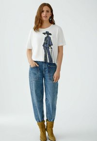 Weißes T-Shirt mit kurzen Ärmeln und blauem Grafikdesign eines Figuren, kombiniert mit lockeren blauen Jeans und gelben Stiefeletten. Einfacher Hintergrund.