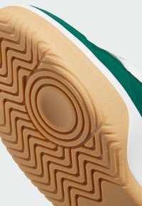 Grön och vit atletisk sko sula med en strukturerad, tan gummiyta som har ett zick-zack mönster och en cirkulär detalj i mitten.