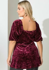Bordowy aksamitny top z bufkami, kwadratowym dekoltem na plecach, podkreśloną talią i aksamitną teksturą. Wyrazisty tatuaż w kształcie serca na plecach.