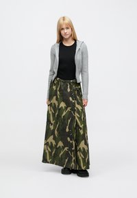 Hall halli lukuga pusa, must särk ja pikk kamuflaažseelik rohe-roheliste ja pruunide mustritega, kinnitatud kahekordsete pandlatega. Mustad platvormkingad.