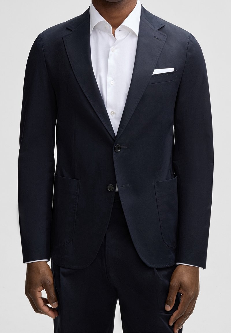 Homme portant un blazer bleu marine foncé sur une chemise blanche, avec un mouchoir blanc visible dans la poche poitrine du blazer.
