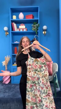 Femme aux longs cheveux blonds tenant une robe fleurie sur un cintre en bois devant un mur bleu avec des étagères et des objets décoratifs.
