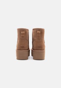 Botas de ante marrón hasta el tobillo con suela de plataforma, que cuentan con un logotipo en la parte trasera, tacón plano y un acabado texturizado. Tiras de agarre sencillas a los lados.
