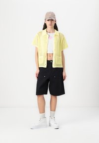 Cardigan giallo a rete aperta con maniche corte sopra un top corto bianco, abbinato a pantaloni shorts neri e sneakers bianche. Un cappellino completa il look.