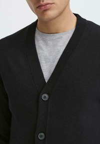 Schwarze Strickjacke mit V-Ausschnitt, mit gerippten Kanten und drei dunklen Knöpfen, getragen über einem hellgrauen T-Shirt. Glatte Textur.