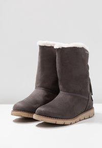 Bottes de cheville en suède gris avec doublure en fausse fourrure en haut, semelle en caoutchouc texturée et design plat. Présente un détail à franges sur le côté.