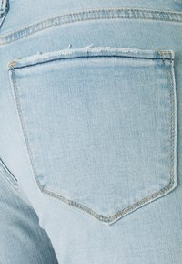 La tela de mezclilla azul claro presenta un solo bolsillo trasero con bordes deshilachados y una costura reforzada a lo largo de la abertura. Acabado suave y texturizado.