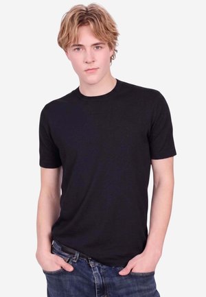 Junger Mann mit hellbraunen Haaren trägt ein schwarzes T-Shirt und blaue Jeans, steht mit den Händen in den Taschen vor einem einfarbigen weißen Hintergrund.