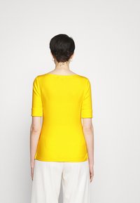 Žlutý top s krátkými rukávy a lodním výstřihem, přiléhavého střihu a hladké textury. Zpětný pohled ukazuje čistý design bez vzorů či ozdob.