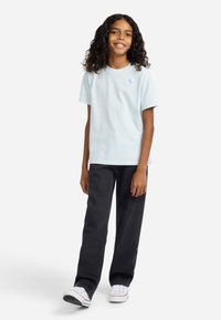 Mlada osoba s dugom kovrčavom kosom, obučena u svijetloplavu majicu, crne traperice i bijele tenisice, stoji i smiješi se.