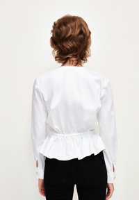 Blusa bianca a maniche lunghe con vita arricciata e dettaglio peplum, dotata di polsini con bottoni e una texture liscia e senza pieghe.