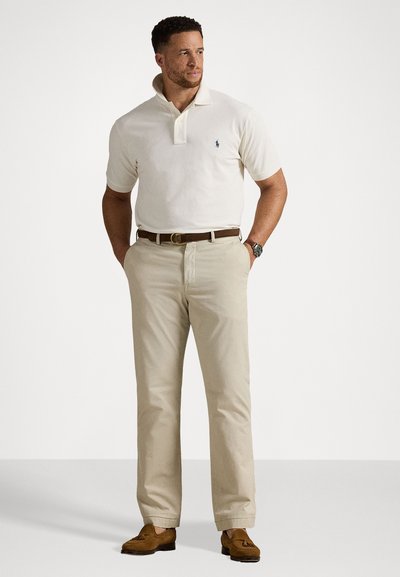 Beige kokvilnas polo kreklis ar apkakli, kopā ar gaiši khaki auduma biksēm un brūnām zamšādām mokasīnēm. Uz krekla ir ievērojams zils logotips.