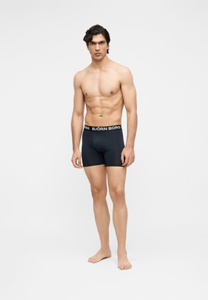 Fit Mann steht barfuß in schwarzen Björn Borg Boxershorts vor einem einfarbigen hellgrauen Hintergrund.