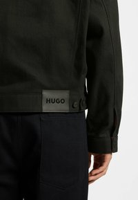 Musta takki teksturoidulla pinnalla, jossa on "HUGO"-logokilpi, napit yksityiskohdissa ja sivuhaarat, yhdistetty tummiin farkkuihin.