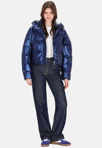 Sötétkék, fényes felületű, rövidített fazonú pufi dzseki kapucnival és nagy első zsebekkel, farmernadrággal és kék sportcipővel párosítva.