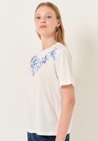 T-shirt bianco a maniche corte realizzato in tessuto morbido, con ricami floreali blu lungo il colletto e le spalle. Taglio semplice e rilassato.