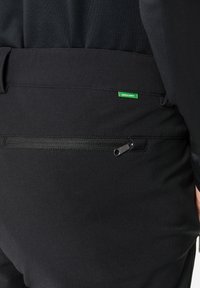 Pantaloni neri con una tasca con zip sul retro, realizzati in un tessuto liscio. Un piccolo tag verde "GREEN SHAPE" è visibile sopra la tasca.