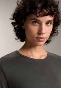 Nuori henkilö, jolla on kihara tummat hiukset, pukeutuneena tummanharmaaseen pyöreäkaulaiseen paitaan, katsoo suoraan kameraan neutraalilla ilmeellä.