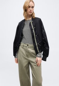 Ξανθιά γυναίκα που φοράει μαύρο μπουφάν τύπου bomber πάνω από μακρυμάνικο ριγέ μπλουζάκι σε άσπρο και μαύρο και ανοιχτόχρωμο μπεζ φαρδύ παντελόνι, μπροστά σε απλό φόντο.