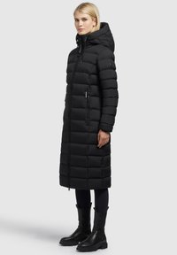 Fekete, hosszú tollkabát kapucnival, steppeléses mintával, cipzáras zsebekkel és elasztikus mandzsettákkal. Fekete bokacsizmával viselve.