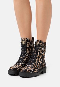 Nogi w czarno-brązowych, lampasowych butach typu lace-up combat z grubymi czarnymi gumowymi podeszwami na białym tle.