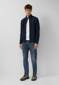 Veste zippée bleu marine avec un tissu texturé, col montant et deux poches latérales, associée à un jean bleu et des bottes grises à lacets.