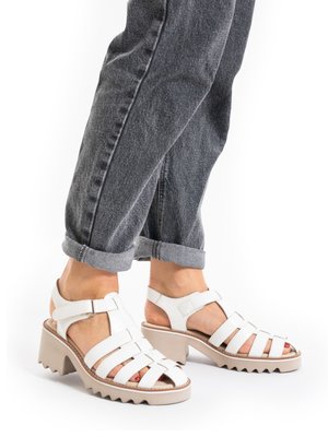 White heeled sandals with multiple straps worn with rolled-up dark gray jeans, showing feet with red nail polish.