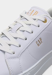 Baskets en cuir blanc avec lacets blancs, œillets dorés et logo "GAP" doré sur la languette et le côté.