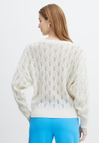 Elfenbeinfarbener gestrickter Pullover mit einem Rautenmuster, offener Webart und geripptem Saum. Die Ärmel sind puffig, und die Rückseite ist leicht länger mit strukturiertem Detail.