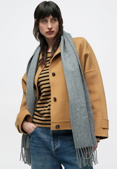 Veste en mélange de laine moutarde avec de gros boutons noirs, écharpe grise et pull rayé noir et or, associée à un jean en denim bleu.