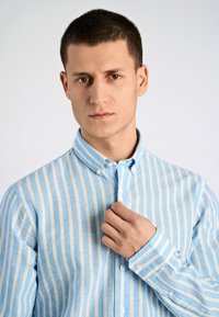 Camicia a righe blu chiaro e crema, in cotone, con colletto a bottoni, che mostra una mano che regola il colletto, texture liscia.