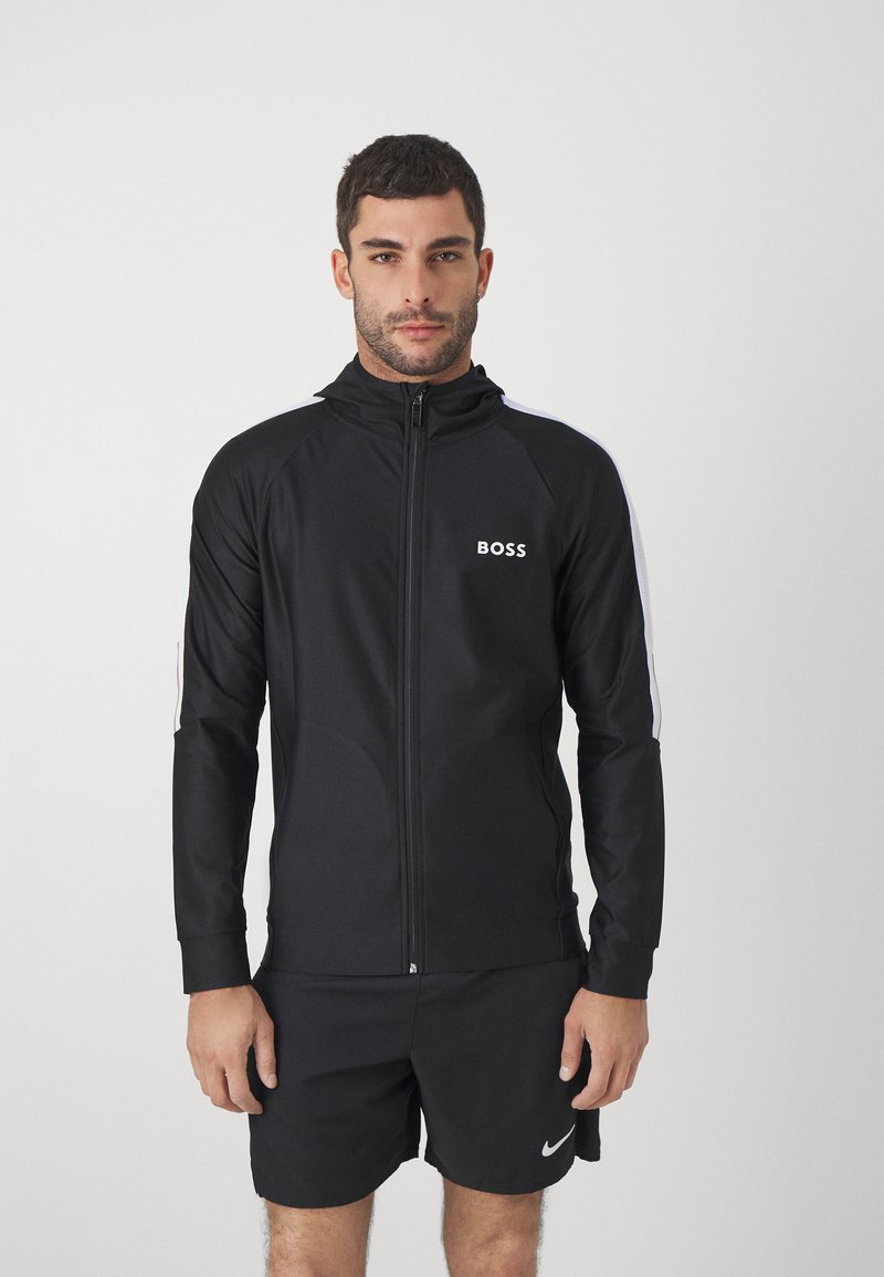 Mann in schwarzer Zip-Jacke mit "BOSS"-Logo und schwarzen Nike-Shorts, vor einem einfarbigen weißen Hintergrund stehend.