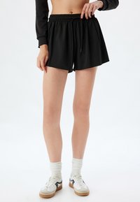 Pantalones cortos deportivos negros con una cintura elástica y cordón ajustable. Tejido suave, corte a la rodilla, combinados con zapatillas blancas y grises.