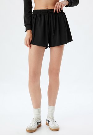 Sorte træningsshorts med elastisk talje og justerbar snor. Glat stof, knælængde, parret med hvide og grå sneakers.