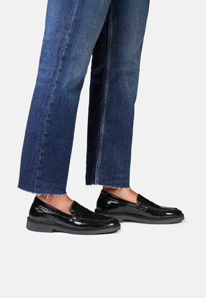 Personne portant un jean denim bleu coupé et des mocassins noirs brillants sans lacets, debout sur un fond blanc.