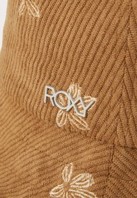 Bruin korduroy stof met ribbelachtige textuur. Bevat witte bloemenborduursels en een zilveren "Roxy" logo accent.