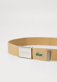 Cinturón de tela beige con un cierre de metal que presenta la grabación "LACOSTE" y un parche con el logo del cocodrilo verde. Superficie texturizada.