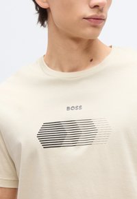 Νεαρό άτομο που φοράει ένα κρεμ T-shirt με μαύρο ριγέ γεωμετρικό σχέδιο και την επιγραφή "BOSS" πάνω στο στήθος, γυρισμένο λίγο στο πλάι.