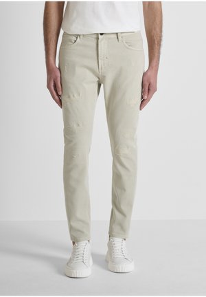 Hombre con jeans ajustados color beige claro con parches desgastados y zapatillas blancas, de pie frente a un fondo blanco liso.