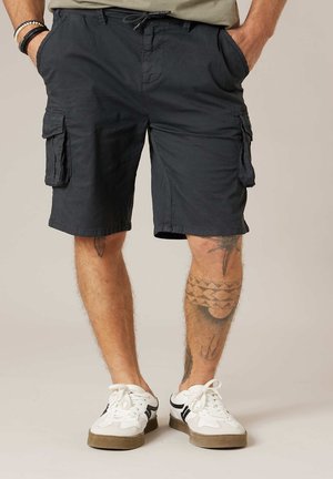 Homme portant un short cargo noir avec des poches latérales, des baskets blanches et des tatouages sur les jambes, debout les mains dans les poches.