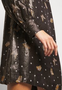 Main et avant-bras à côté d'une robe métallique sombre avec motif papillons et pois blancs, manches longues avec poignets et tissu fluide.