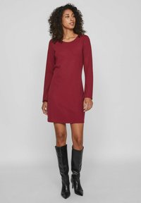 Vestido de manga larga burdeos con cuello redondo, ajuste ceñido y tejido suave, combinado con botas negras de tacón puntiagudo hasta la rodilla.