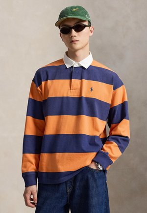 Ung mand iført grøn kasket, sorte solbriller, rugbytrøje med orange og marineblå striber og blå jeans med den ene hånd i lommen.