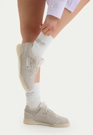 Person trägt hellbeige Turnschuhe und weiße Reebok-Socken, zieht die Socke an einem Bein vor einem schlichten weißen Hintergrund zurecht.