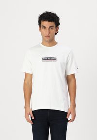 Valge puuvillane T-särk ümarate kaelustega, millel on must ristkülikukujuline logo, mille keskel on valge ja sinise aktsendiga tekst "Tommy HILFIGER".