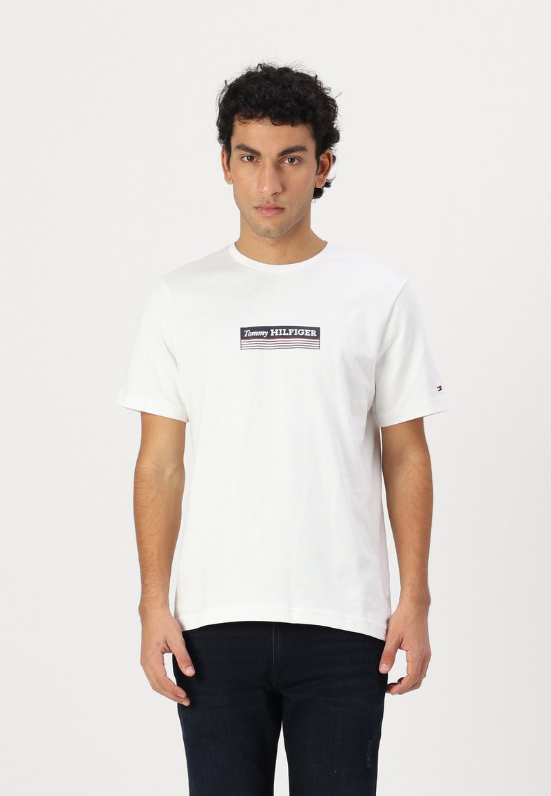 Valge puuvillane T-särk ümarate kaelustega, millel on must ristkülikukujuline logo, mille keskel on valge ja sinise aktsendiga tekst "Tommy HILFIGER".